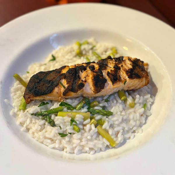Grilled salmon with a dark, crispy skin, served over creamy white risotto with green asparagus pieces.