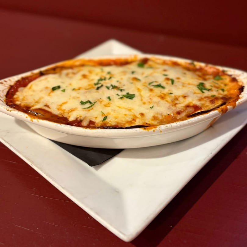 Baked lasagna in a white oval dish, topped with melted cheese and parsley, on a white square plate.