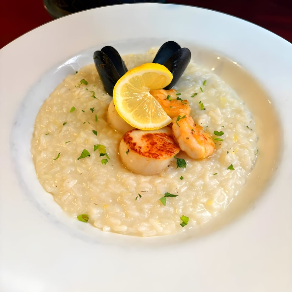 Creamy seafood risotto with mussels, scallops, shrimp, parsley, and a lemon slice in a white bowl.